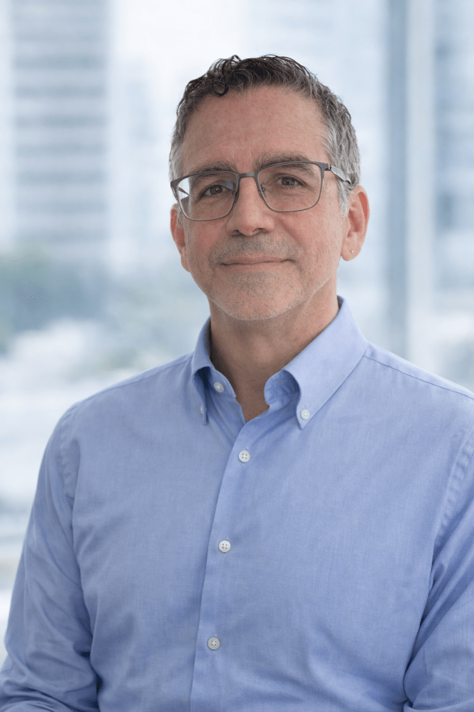 A middle-aged man with glasses, smiling and wearing a light blue button-up shirt, standing in front of a blurred city background.
